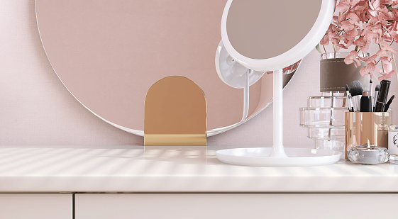 Beautiful and luxury wooden white dressing table with a vase of pink flower and frameless round mirror in bedroom with sunlight from window blinds on wallpaper wall for product display