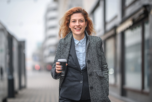 Attractive business woman on the street in a city, having her morning coffee and dashing to work, smiling. Looking at camera.