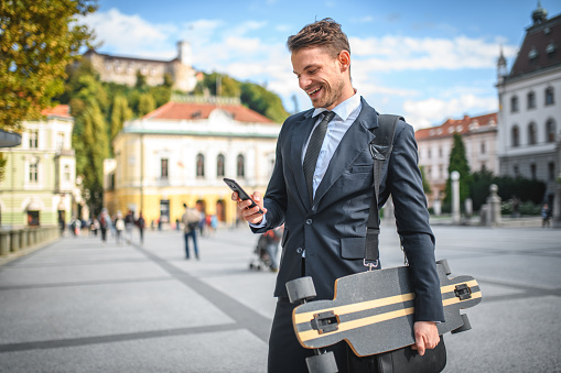 3/4 side shot of a young male executive on his way to the office, wearing a business suit and tie, carrying a longboard and bag, holding a smart phone in hand, reading mails with a broad smile. Walking in old town center next to a park. Looking away.