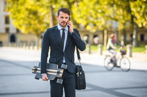 Busy male entrepreneur, holding a longboard and carrying a messenger bag, talking on his smart phone, walking down the street of old town, big park behind him, dressed for success. Early morning, slowly going to work. Focus on foreground. Looking away. 3/4 shot.