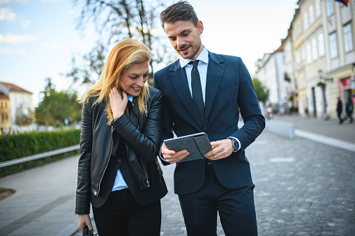 Attractive female and male business colleagues on their way to a meeting, man showing something on his digital tablet and talking, woman carrying a soft briefcase, both dressed elegantly. 3/4 shot, looking away, old town in background.