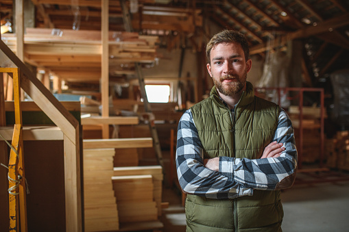 Portrait of a Caucasian young adult carpenter standing in a workshop with his hands folded looking at camera.