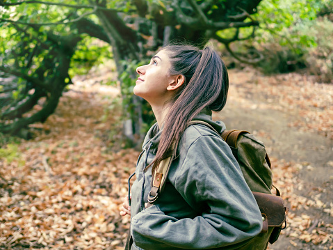 Side view of woman with backpack in the forest admiring the beauty of wild nature, looking up with a happy and satisfied smile.