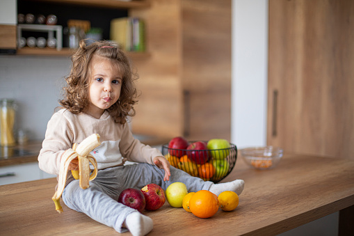 Cute baby girl eating fruits and a banana in domestic kitchen at home