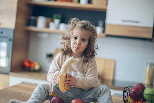 Cute baby girl eating fruits and a banana in domestic kitchen at home