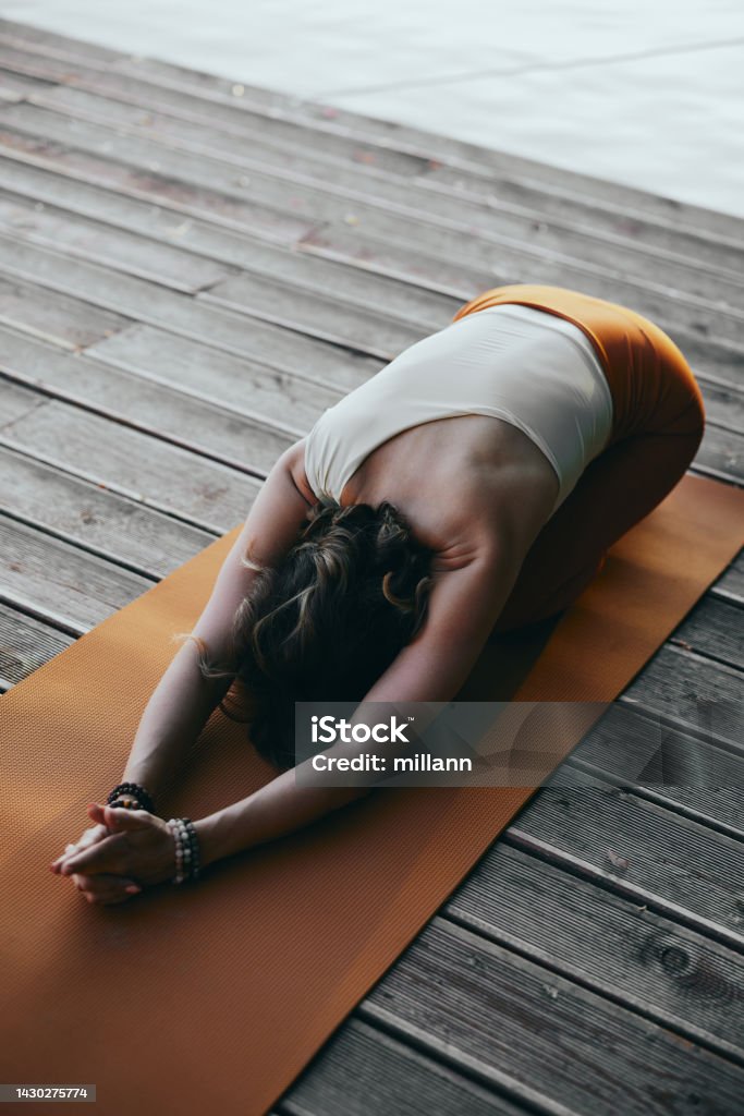 A yogi woman practices yoga on a dock. She is in a balasana yoga posture. Yoga Stock Photo