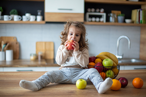 Cute baby girl eating fruits and an apple in domestic kitchen at home