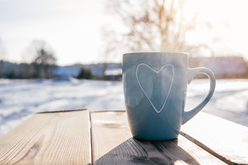 Turquoise blue cup with heart rising steam on wooden table against backdrop of trees and houses with snow on cold winter day. Hot warm drink in frosty weather
