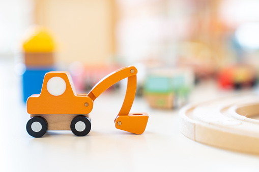 An orange wooden toy excavator is placed on the table in front of other blurry wooden toys.