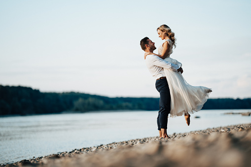 Romantic wedding couple barefoot on pebblestone beach at river on late summer afternoon, groom carrying bride, smiling at each other, full length, copy space