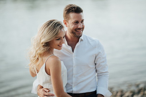 Medium shot portrait smiling couple outdoors in summer at river, background blurred, woman with long hair and white dress, man in shirt
