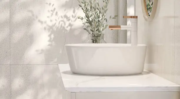 Modern and minimal design of cream colored bathroom vanity with marble counter top and white round ceramic washbasin with vase of houseplant in sunlight from window on granite tile wall for personal care and toiletries product display