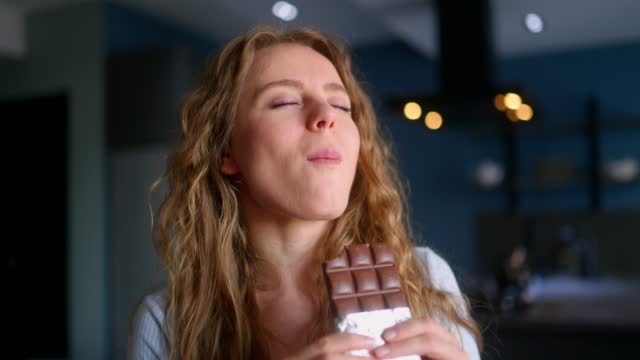 Hungry woman bites into a bar of chocolate and enjoys dessert. Curly blonde girl overeats unhealthy sweet food. Female stuffs her face into candy at kitchen interior. Healthy diet, overweight concept.