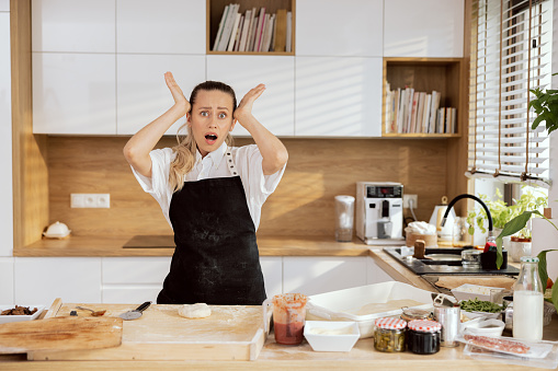 Confused shocked young woman lady chef wearing in apron holding hands near head looking at camera opeping mouth. Pizza ingredients on table tomato sauce olives asparagus butter flour eggs baking.