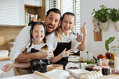 Funny family in aprons looking at camera smiling. Prepared pizza ingredients on wooden surface. Happy kid spending time with parents in modern kitchen at weekend preparing dinner