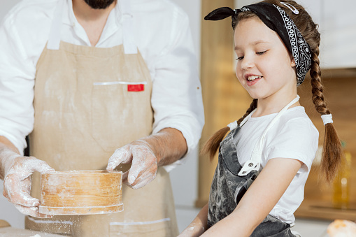Close-up shot smiling daughter in apron helping father sieving flour preparing cooking baking homemade pizza pasta gnocchi cookies biscuits. Father's hands in background.
