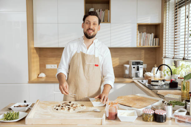 istock Good-looking man father husband wearing apron looking at camera smiling while dressing homemade dough for pizza with kitchen ingredients mushrooms salami feta 1430318377
