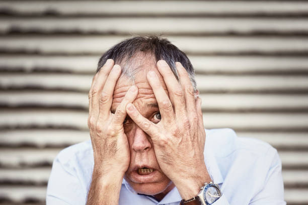 hombre aterrorizado de mediana edad se asoma cautelosamente a través de sus dedos, lleno de consternación - nail biting guilt men anxiety fotografías e imágenes de stock