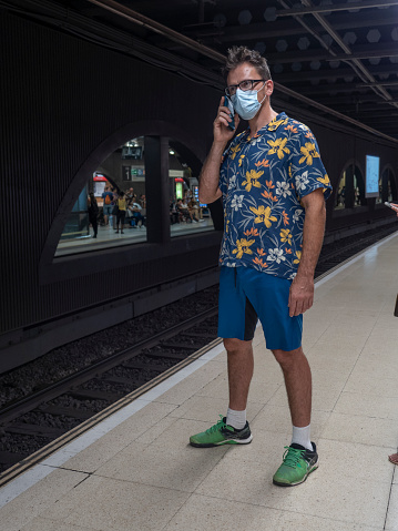 man in cheerful clothes talking on a mobile phone inside a city underground station, wearing a protective mask and waiting for the underground.
