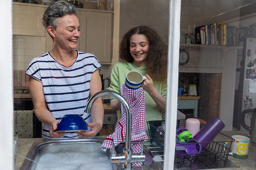 Waist up shot of a mature female mother and daughter washing the dishes together. They are smiling and laughing together. The photo is shot through a wiondow.
