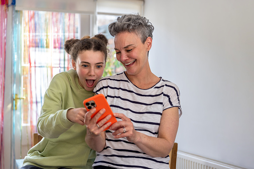 Waist up shot of mother and daughter talking in the kitchen. The mother is holding\showing her daughter something on an orange mobile phone. The daughter has her mouth open in amazement.