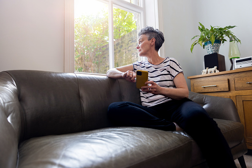 A three quater length shot of a mature female adult sitting on a leather sofa. She has her phone in her hands whilst looking out the window.
