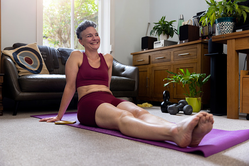 Full shot of mature adult sitting with her legs extended out on an exercise mat smiling, looking away from the camera. She has her hands behind her back wearing gym gear.