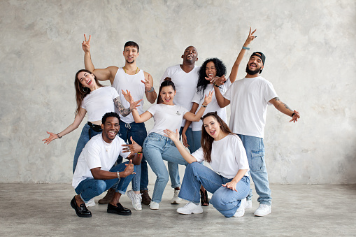 Multiethnic group of happy friends smile and raise hands up. Diverse young people standing together in white shirts and jeans on background wall in studio