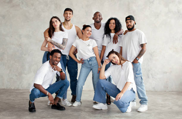 Multiethnic group of happy friends. Diverse young people standing together in white shirts and jeans on background wall in studio stock photo