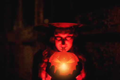 spooky witch holding glowing pumpkin and looking at camera on halloween portrait