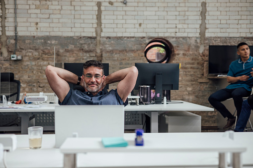 A businessman sitting at his desk in a modern office in Hexham, Northumberland. He is sitting back with his hands behind his head while he looks at his laptop and smiles. One of his colleagues is in the background.