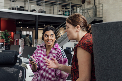 Two businesswomen standing in a modern office discussing new business ideas in Hexham, Northumberland. One woman is showing her colleague something on her mobile phone.