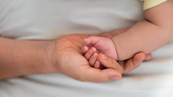 Young mother holding baby hand and showing finger and fingernail on white sheet bed. Closeup of baby sleeping on the mother's room. Family and home concept