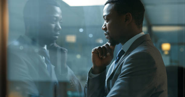 pensiero, idea e mentalità con un uomo d'affari in piedi nel suo ufficio di notte mentre è concentrato sul futuro della sua azienda. missione, visione e sviluppo con un dipendente maschio al lavoro da una finestra - reflective glass foto e immagini stock