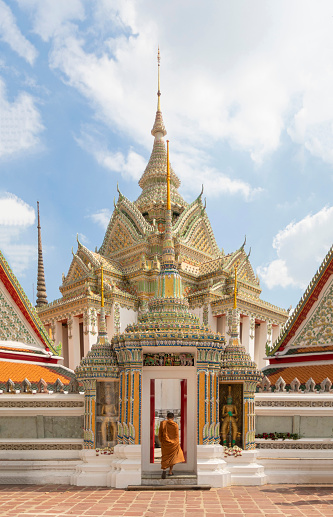 A wide shot view of a religious Buddhist monk wearing a traditional ape walking into the ancient temple Wat Phra Chetuphon or Wat Pho, with golden and shiny patterned architecture exterior, through the small entrance with two old Buddhist statues on the left and right of the door, in Thailand Bangkok in a Sunny day.