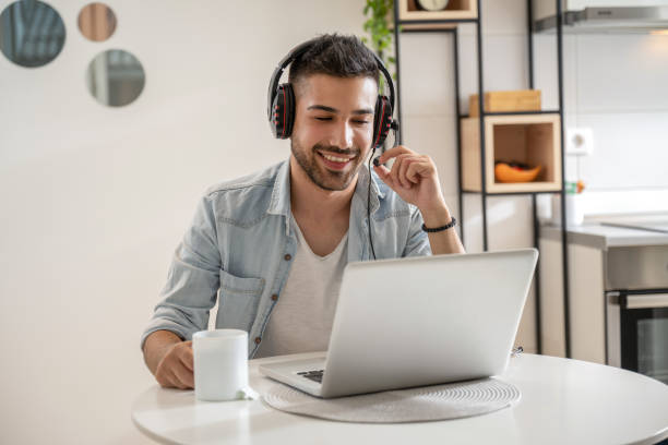 Young man working as agent at call center from home while drinking coffee. Young friendly man in good mood working as agent at call center from home while drinking coffee. Wearing headset and using laptop. working at home stock pictures, royalty-free photos & images