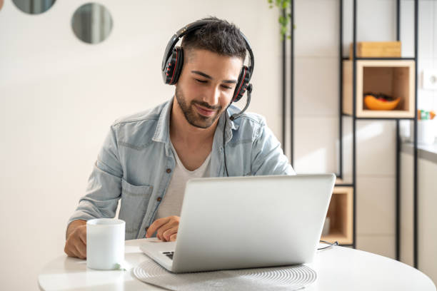istock Young friendly man working as agent at call center from home while drinking coffee. Wearing headset and using laptop. 1430348739