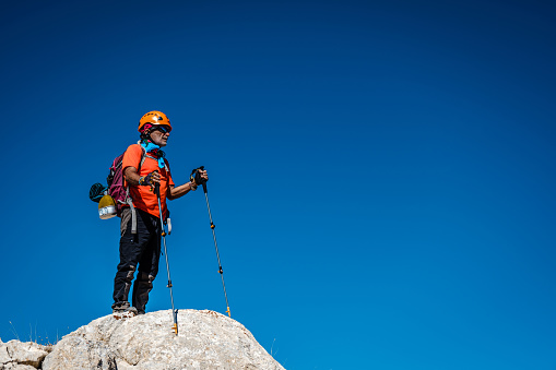Man aged 60-65. Hiker in orange t-shirt, helmet, back, walking pole, sunglasses. He is climbing the mountain. High altitude. High adventure spirit. Clear blue skies. High altitudes, mountainous extreme terrain. Full size model