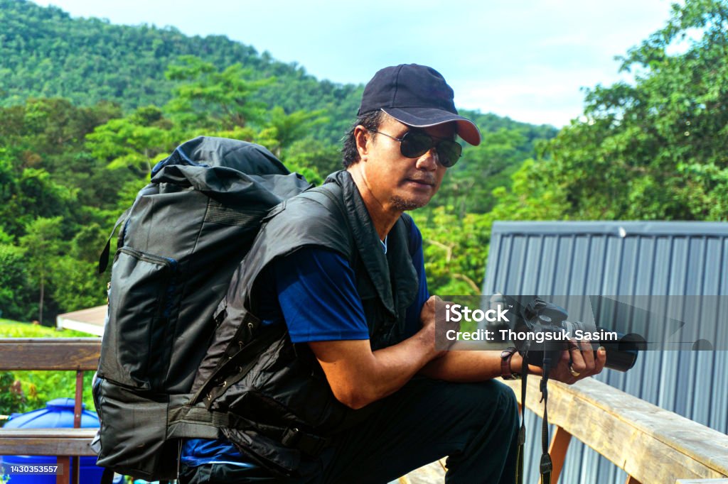 Backpackers capture the beauty of nature standing on a hillside wooden terrace. Male tourists travel to capture the beauty of nature with photographic equipment standing on a tropical wooden balcony. The concept of photographing mountains of dark-skinned Thai people Capital Region Stock Photo