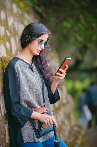 A beautiful Asian/Indian young woman in sunglasses uses a smartphone while standing close to a stone wall by the side of an allay.