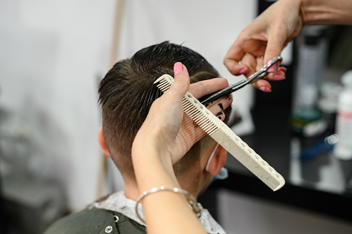 A teenager in a beauty salon gets a haircut, a hairdresser cuts a teenage boy's hair, a haircut with scissors and a comb.