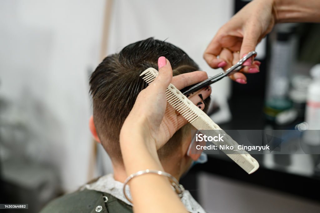 A teenager in a beauty salon gets a haircut, a hairdresser cuts a teenage boy's hair. A teenager in a beauty salon gets a haircut, a hairdresser cuts a teenage boy's hair, a haircut with scissors and a comb. Cutting Hair Stock Photo