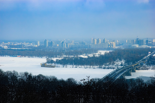 Panoramic view of the Kiev's left bank during a foggy winter day. The Dnieper river is frozen. Trees, road, bridge and buildings  appear in the distance