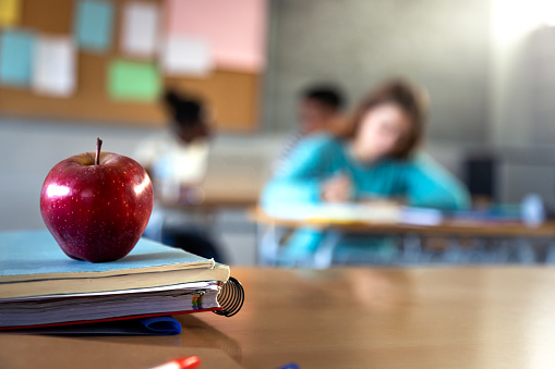 Apple and a pile of books and pens on teacher table in classroom. Blurred background scene of high school student doing homework. Focus on foreground. Copy space. Education concept.