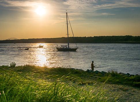 A man fishes along the the banks of the Cape Cod Canal as a yacht passes and an August sun slowly sets.