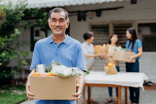 Image of a senior Asian Chinese volunteer holding a box of donated groceries at food bank. Volunteer ready to give out a box of free food and groceries to the people impacted by financial difficulty.