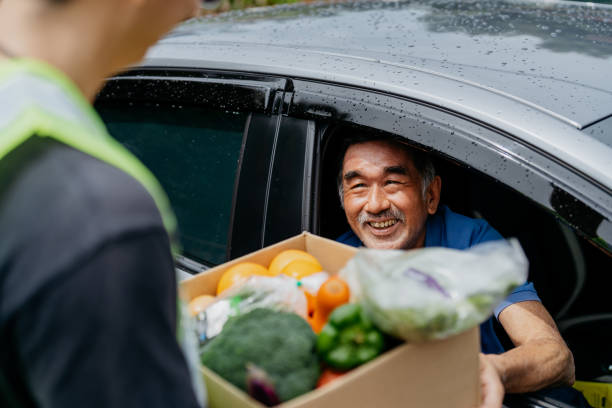 Asian senior man receiving donated groceries from volunteer at drive through food bank Image of an Asian Chinese senior man receiving a box of free food and groceries from volunteer at a drive through food bank food bank stock pictures, royalty-free photos & images