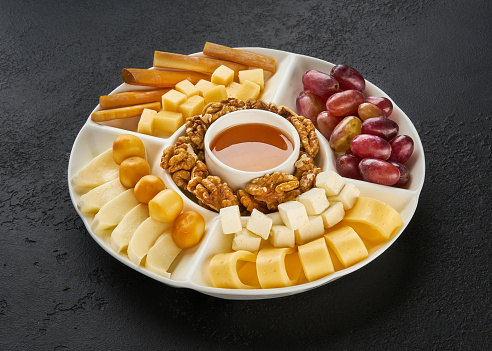 cheese plate served with grapes, nuts and honey on a black wooden background