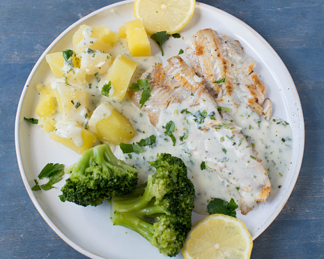 Plate with homemade fish fillet, boiled potatoes, broccoli and herb, bechamel sauce. Served isolated on blue wooden table. Top view and closeup