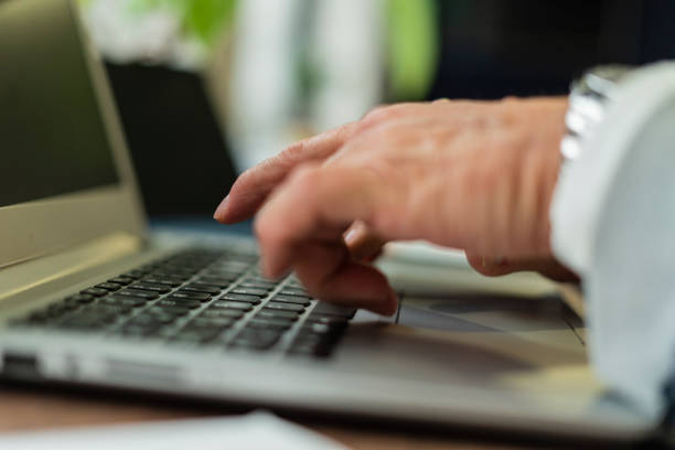 hands of an older man in his office dressed formally working with his electronic media that he has on his desk and documents that relate to his work